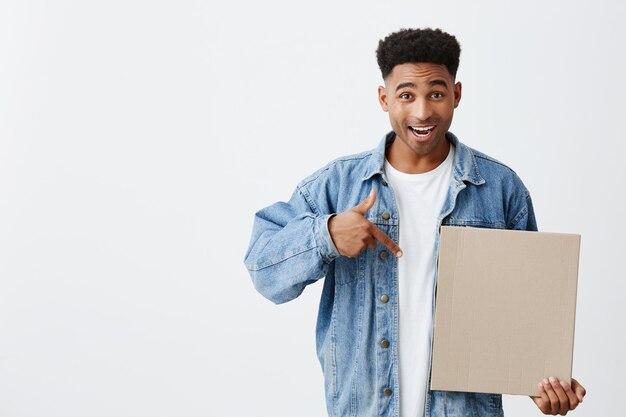 OMG. Attention. Isolated on white portrait of young attractive black-skinned man with afro hairstyle in white tshirt and denim jacket holding cardboard in hand, pointing with happy expression