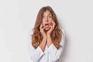 Free photo omg concept speechless scared beautiful european woman with long hair keeps mouth opened stares bugged eyes reacts on shocking news keeps hands on face dressed casually isolated over white background