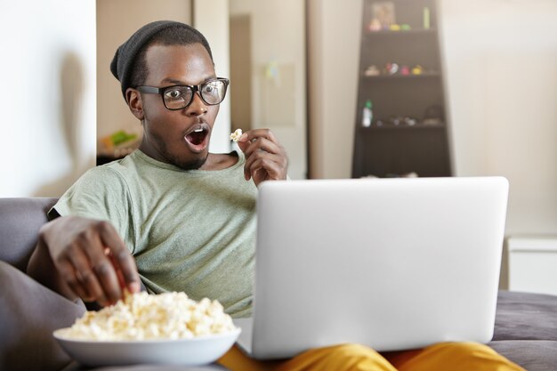 Omg. Excited emotional young dark-skinned man in hat and rectangular glasses sitting on couch at home with laptop computer and bowl of popcorn, watching detective series online with mouth wide open