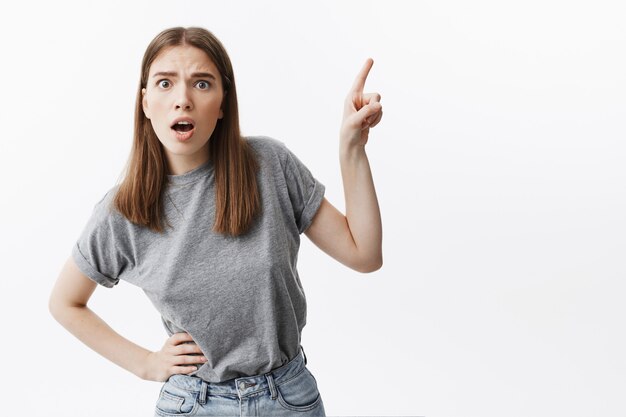 OMG. Funny young attractive european student girl with brown hair in casual stylish clothes  with iopened mouth and shocked expression, pointing with index finger on free space on wall