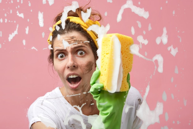 Free photo omg. headshot of emotional housewife with dirty face washing window using sponge and chemicals, feeling shocked as she has to do all cleaning by herself, looking, keeping mouth wide opened