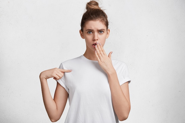 Omg, look there! Shocked woman in bewilderment covers mouth, wears casual white t shirt, points with fore finger at blank copy space, isolated over white background. People and advertisment concept