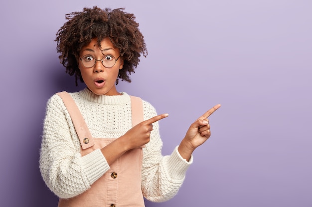 Omg, look at this. Nervous surprised Afro American woman points aside, demonstrates something on blank space, has bugged eyes, wears round spectacles for good vision, dungarees. Promotion concept