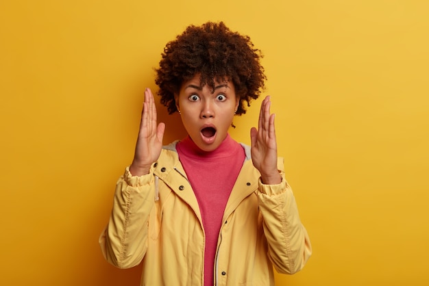 Omg, so big!  portrait of excited curly haired shocked woman shows huge size, raises palms and measures large item, keeps mouth opened, dressed in casual anorak, poses over yellow wall