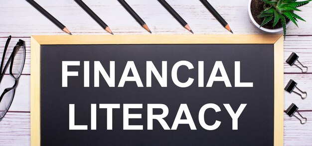 На светлом деревянном столе доска с текстом финансовая грамотность карандаши растения очки и скрепки бизнес-концепция