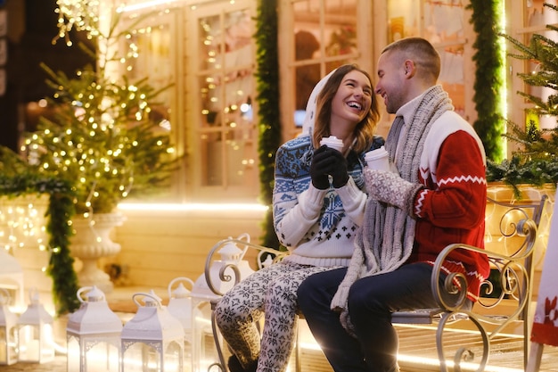 크리스마스 이브에 로맨틱한 두 사람의 매력적인 커플이 벤치에 앉아 따뜻한 음료를 즐기고 있습니다.