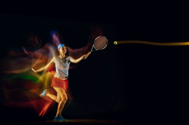 Одна кавказская женщина, играющая в теннис, изолирована на черной стене в смешанном и светлом свете. Подходит молодой игрок женского пола в движении или действии во время спортивной игры. Понятие движения, спорта, здорового образа жизни.