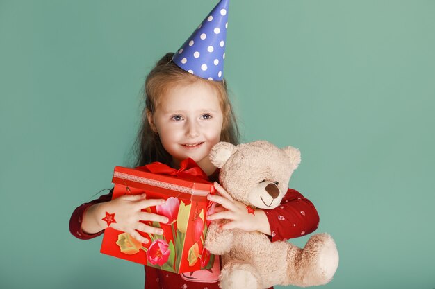 One funny happy child with present and bear toy dressed in birthday hat on the green background, sincerely smiling