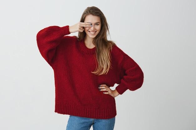 One glance and you do not have a chance. Emotive good-looking girl in red loose sweater covering eye with gun gesture, smiling broadly and flirting, looking sensually  .