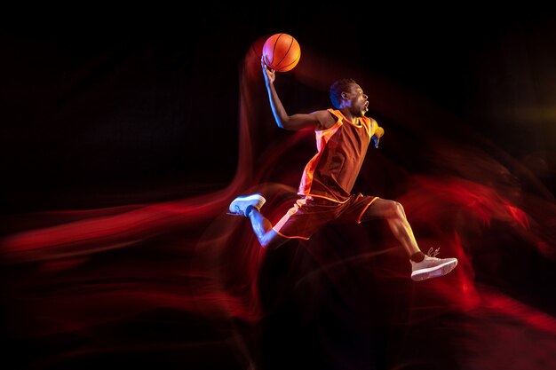 One jump before the winning. African-american young basketball player of red team in action and neon lights over dark studio background. Concept of sport, movement, energy, dynamic, healthy lifestyle.