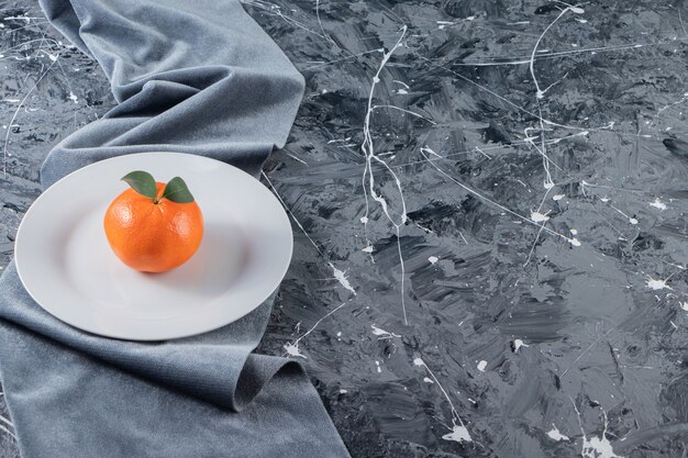 One tangerine on a plate on a pieces of fabric , on the mixed table.