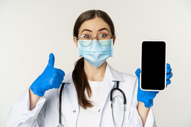 Free photo online medical help concept enthusiastic young woman doctor in face mask showing thumbs up and mobil...