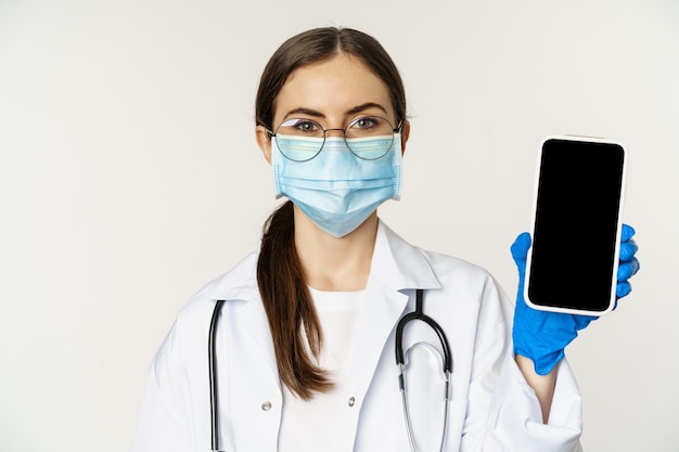オンライン医療ヘルプの概念。眼鏡とフェイスマスクの女医、白い背景の上に立って、患者のための携帯電話の画面、アプリのインターフェースまたはウェブサイトを表示