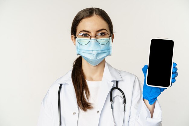 Online medical help concept. Woman doctor in glasses and face mask, showing mobile phone screen, app interface or website for patients, standing over white background