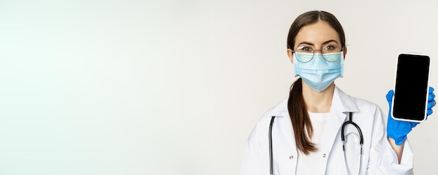 Free photo online medical help concept woman doctor in glasses and face mask showing mobile phone screen app in