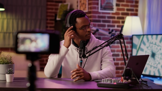 Online radio host answering internet call from listener while recording vlog using digital video camera in home podcasting studio. African american podcaster talking to audience using microphone.