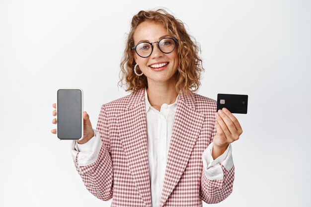 Online shopping and business. Young female entrepreneur showing mobile phone screen with credit card and smiling, standing over white background.