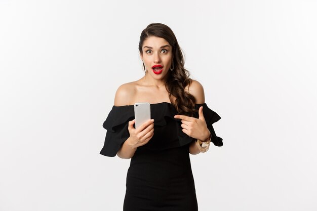 Концепция покупок в Интернете. Стильная женщина в черном платье, макияж, указывая пальцем на мобильный телефон с удивленными эмоциями, стоя на белом фоне.