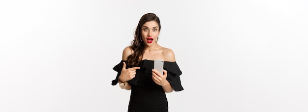 Online shopping concept stylish woman in black dress wearing makeup pointing finger at mobile phone