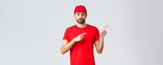 검역 및 테이크아웃 개념 동안 온라인 쇼핑 배달 빨간색 유니폼 모자와 티셔츠 웃는 카메라를 입은 자신감 있는 수염 택배는 고객에게 올바른 방향을 가리키는 손가락을 제공합니다.