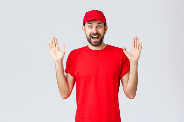 온라인 쇼핑, 검역 중 배달 및 테이크아웃 개념. 빨간 티셔츠와 모자, 회사 유니폼을 입은 행복한 쾌활한 택배, 회색 배경에 서 있는 놀라움과 즐거운 손