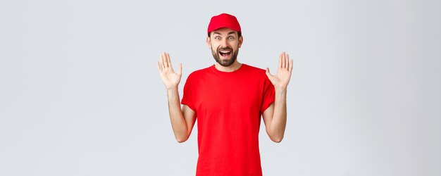 検疫と持ち帰りの概念の間にオンラインショッピングの配達赤いTシャツとキャップ会社の制服を着た幸せな陽気な宅配便は驚いて面白がって立っている灰色の背景を手渡します