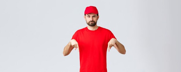 Online shopping delivery during quarantine and takeaway concept Skeptical or suspicious courier in red uniform cap and tshirt pointing fingers down standing grey background