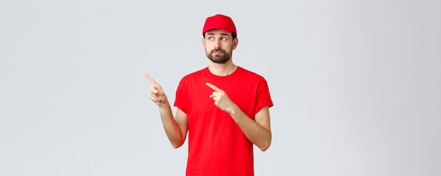 검역 및 테이크아웃 개념 동안 온라인 쇼핑 배달 빨간색 티셔츠와 모자를 쓴 불확실하고 꺼려하는 택배 직원은 왼쪽 상단 모서리를 가리키는 손가락을 실망스럽게 웃었습니다.