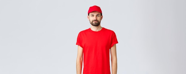 검역 및 테이크아웃 개념 동안 온라인 쇼핑 배달 모자와 티셔츠를 입고 주문을 기다리는 카메라를 바라보는 빨간 제복을 입은 젊은 수염 택배
