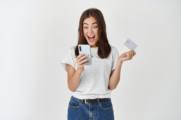 Интернет-магазины и электронная коммерция Счастливая молодая женщина, использующая мобильный телефон и держащая пластиковую кредитную карту, совершая покупку, стоя на белом фоне