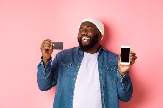 Online shopping. Happy african-american man in beanie laughing, showing credit card and mobile phone screen, standing over pink background