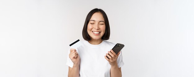 オンラインショッピングクレジットカードとスマートフォンアプリを使用して携帯電話の白い背景を介してウェブサイトで支払う幸せなアジアの女性