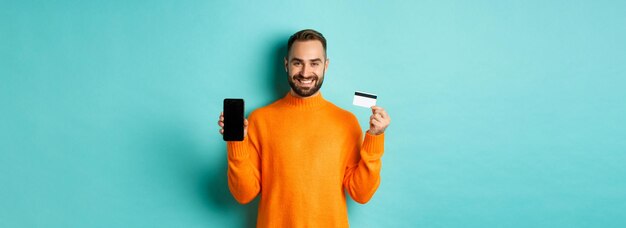 Free photo online shopping happy attractive guy showing mobile phone screen and credit card smiling satisfied s