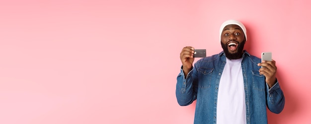 온라인 쇼핑 행복한 수염을 기른 아프리카계 미국인 남자가 신용 카드를 보여주고 구매를 하고 웃고 있습니다.
