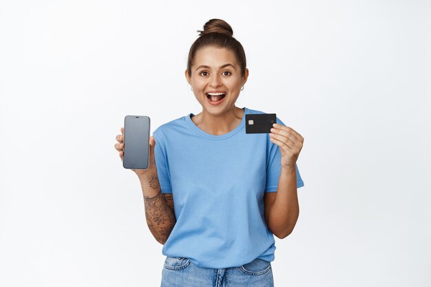 Online shopping. Happy young woman showing credit card and mobile phone empty screen, app interface, standing on white in blue tshirt