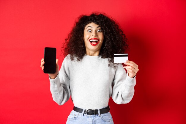 Онлайн покупки. Счастливая молодая женщина, показывающая пластиковую кредитную карту и пустой экран телефона, объявляет о промо-предложении, стоя на красном фоне