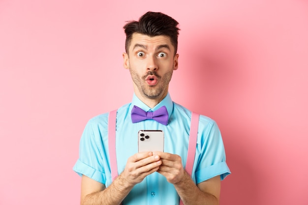Online shopping intrigued guy in bowtie checking out internet promo offer on mobile phone say wow at...