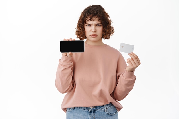 Online shopping and mobile banking. Frowning angry girl showing horizontal smartphone screen and credit card, standing in casual clothes against white background