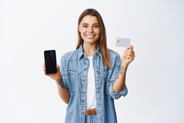 Online shopping Portrait of smiling blond girl recommending shop showing empty smartphone screen and plastic credit card standing over white background