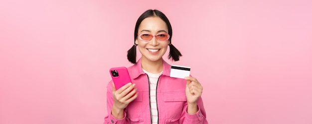 Online shopping Smiling asian girl shopper holding smartphone and credit card paying in mobile app standing over pink background