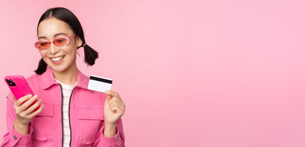온라인 쇼핑 분홍색 배경 위에 서 있는 모바일 앱에서 스마트폰과 신용카드를 들고 웃고 있는 아시아 소녀 쇼핑객
