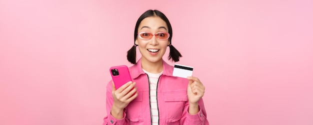 Online shopping Smiling asian girl shopper holding smartphone and credit card paying in mobile app standing over pink background