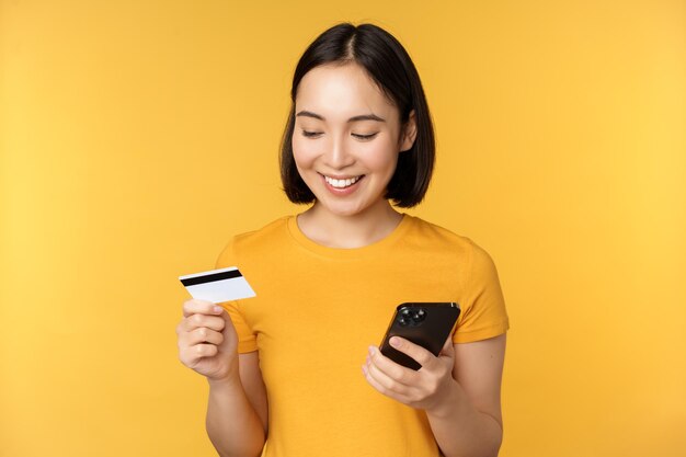 Online shopping Smiling asian girl using credit card and mobile phone app paying contactless order on smartphone application standing over yellow background