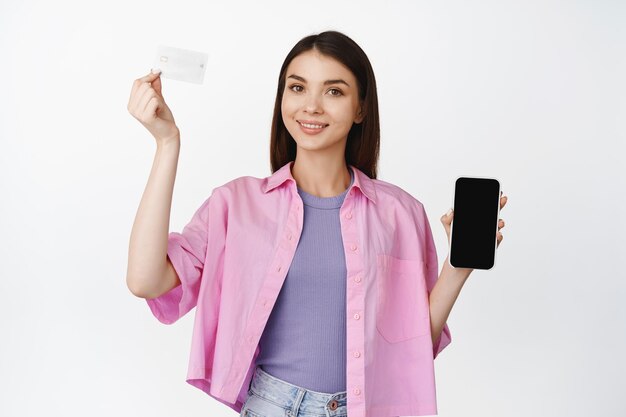 Интернет-магазин Улыбающаяся брюнетка поднимает руку с кредитной картой, показывая мобильный интерфейс экрана смартфона, стоящий на белом фоне
