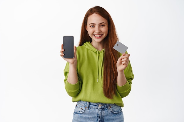 Online shopping. Smiling girl shows mobile app interface and credit card, shops in internet store, buying contactless from home, standing against white background.