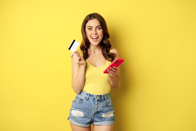 온라인 쇼핑. 스마트폰과 신용 카드를 들고 있는 세련된 브루네트 여성, 앱 결제, 휴대폰 애플리케이션 사용, smth 구매, 노란색 배경 위에 서 있습니다.