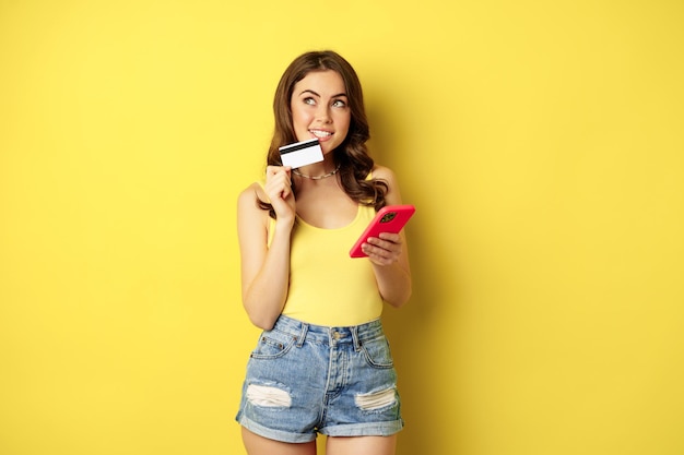 온라인 쇼핑. 스마트폰과 신용 카드를 들고 있는 세련된 브루네트 여성, 앱 결제, 휴대폰 애플리케이션 사용, smth 구매, 노란색 배경 위에 서 있는