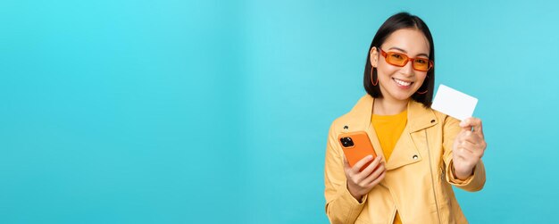Интернет-магазины Стильная молодая азиатка в солнцезащитных очках показывает кредитную карту и с помощью смартфона оплачивает покупку в Интернете, стоя на синем фоне