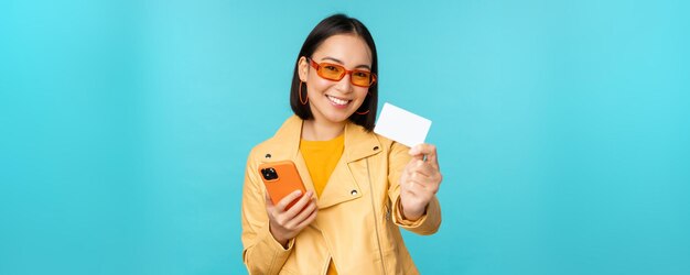 Online shopping Stylish young asian woman in sunglasses showing credit card and using smartphone paying in internet making purchase standing over blue background