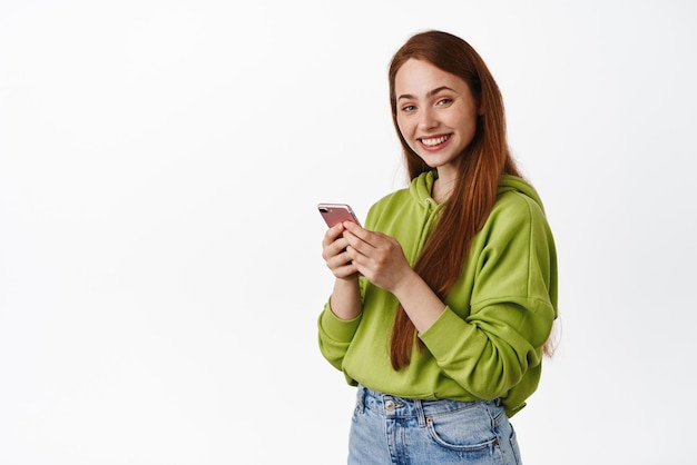 Foto gratuita acquisti online e tecnologia ragazza sorridente dello zenzero che chatta sul telefono guardando felice la fotocamera e l'app di controllo della messaggistica su sfondo bianco dello smartphone